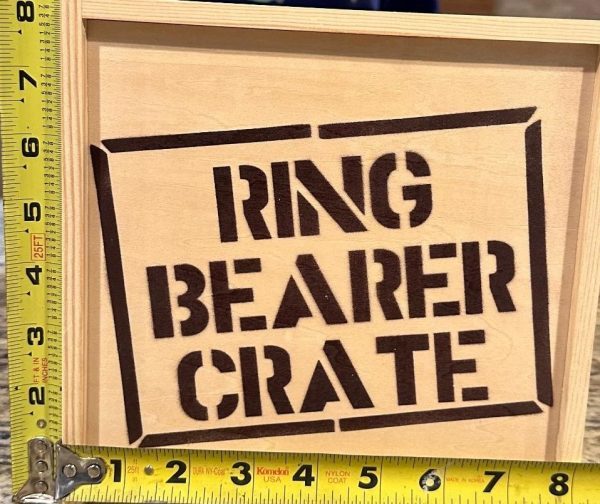 A "Ring Bearer Crate - Wooden Gift Box" with the name stenciled on the front is being measured with two yellow tape measures, one for height and the other for width.